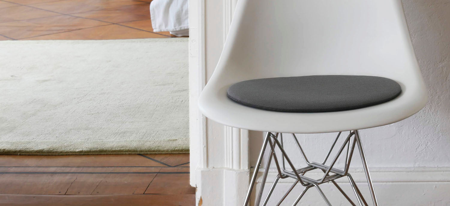 Das foto zeigt einen weissen eames plastic side chair dsr auf dem eine runde sitzauflage der firma discus liegt. Die farbe der sitzauflage ist grau. Der stuhl steht in einem wohnzimmer an der wand.