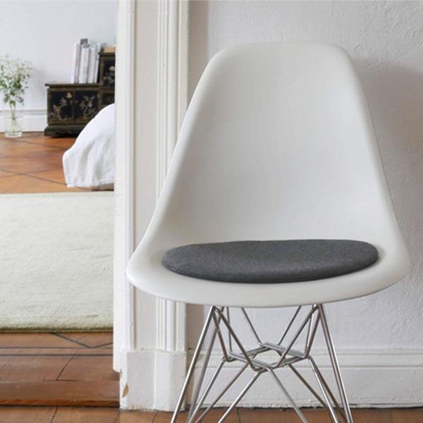 Das foto zeigt einen weissen Eames Plastic Side Chair DSR auf dem eine runde sitzauflage der firma discus liegt. Die Farbe der Sitzauflage ist grau meliert. Der Stuhl steht in einem Wohnzimmer an der Wand.