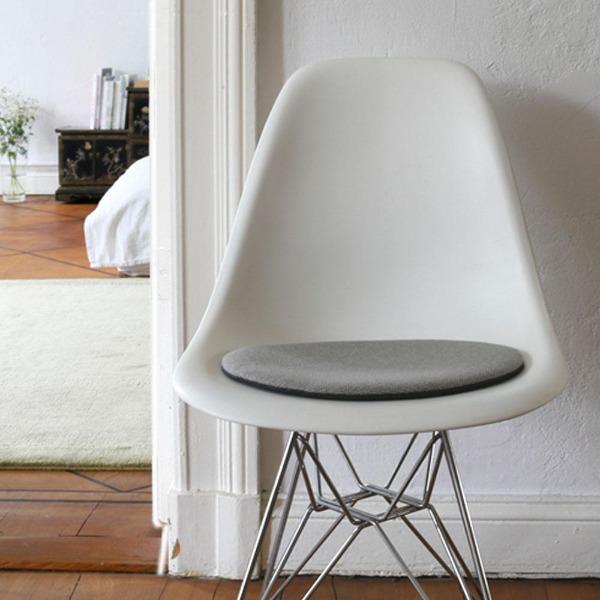 Das foto zeigt einen weissen eames plastic side chair dsr auf dem eine runde sitzauflage der firma discus liegt. Die farbe der sitzauflage ist hellgrau-grau meliert. Der stuhl steht in einem wohnzimmer an der wand.