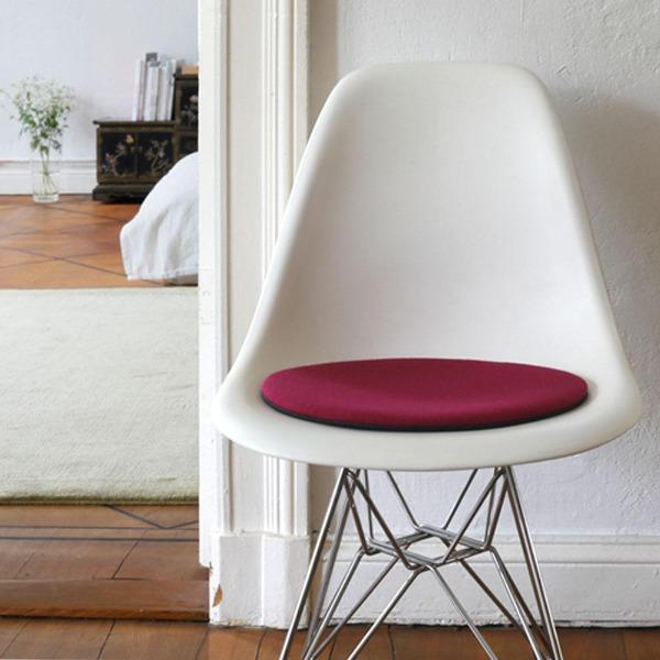 Das foto zeigt einen weissen eames plastic side chair dsr auf dem eine runde sitzauflage der firma discus liegt. Die farbe der sitzauflage ist violett. Der stuhl steht in einem wohnzimmer an der wand.