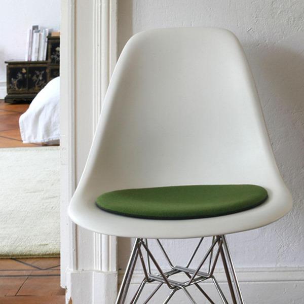 Das foto zeigt einen weissen eames plastic side chair dsr auf dem eine runde sitzauflage der firma discus liegt. Die farbe der sitzauflage ist gruen mit einem leichten gelbstich. Der stuhl steht in einem wohnzimmer an der wand.