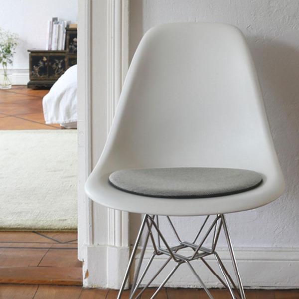 Das foto zeigt einen weissen eames plastic side chair dsr auf dem eine runde sitzauflage der firma discus liegt. Die farbe der sitzauflage ist schwarz/weiß meliert. Der stuhl steht in einem wohnzimmer an der wand.