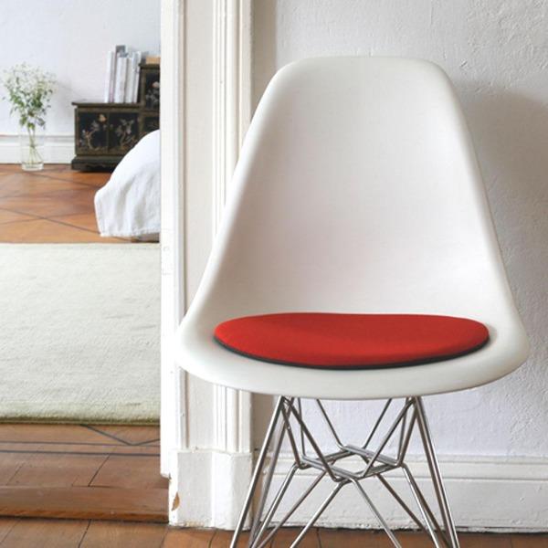 Das foto zeigt einen weissen eames plastic side chair dsr auf dem eine runde sitzauflage der firma discus liegt. Die farbe der sitzauflage ist rot meliert. Der stuhl steht in einem wohnzimmer an der wand.