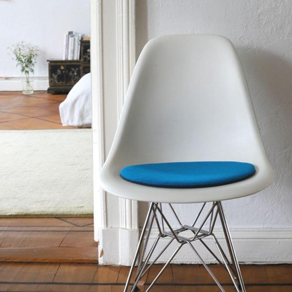 Das foto zeigt einen weissen eames plastic side chair dsr auf dem eine runde sitzauflage der firma discus liegt. Die farbe der sitzauflage ist türkis-blau meliert. Der stuhl steht in einem wohnzimmer an der wand.