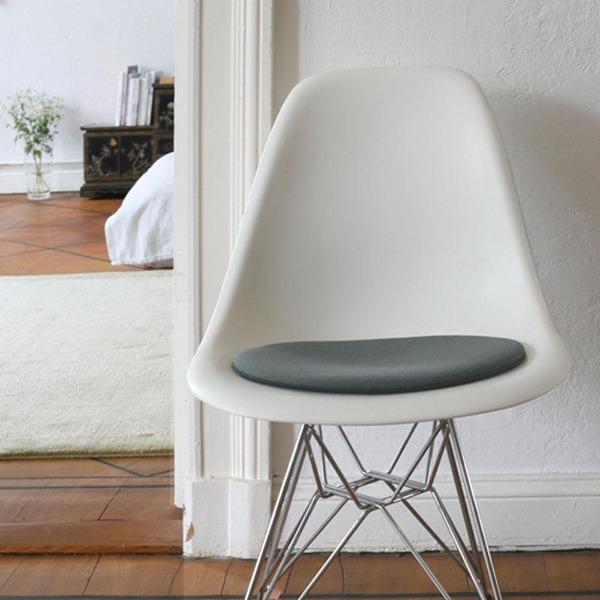 Das foto zeigt einen weissen eames plastic side chair dsr auf dem eine runde sitzauflage der firma discus liegt. Die farbe der sitzauflage ist anthrazit meliert mit einem leichten grünstich. Der stuhl steht in einem wohnzimmer an der wand.