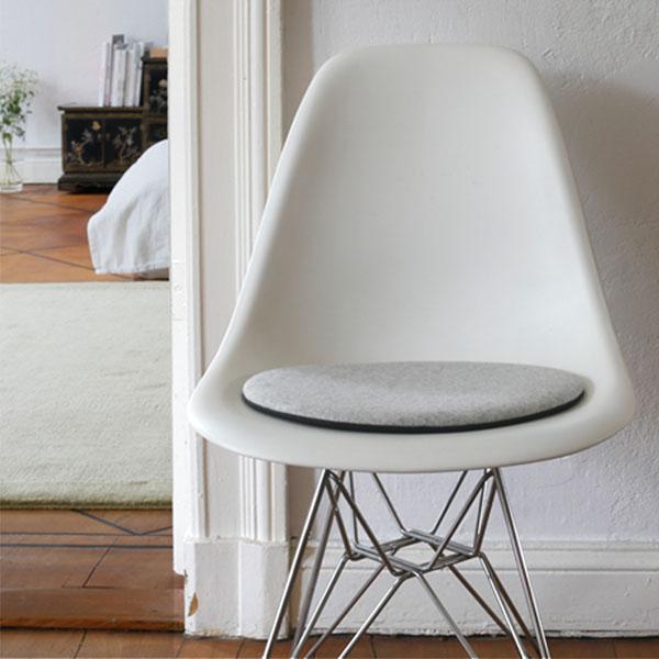 Das foto zeigt einen weissen Eames Plastic Side Chair DSR auf dem eine runde sitzauflage der firma discus liegt. Die Farbe der Sitzauflage ist hellgrau meliert. Der Stuhl steht in einem Wohnzimmer an der Wand.