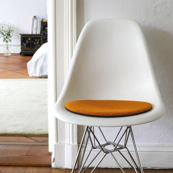 Das foto zeigt einen weissen eames plastic side chair dsr auf dem eine runde sitzauflage der firma discus liegt. Die farbe der sitzauflage ist orange meliert. Der stuhl steht in einem wohnzimmer an der wand.