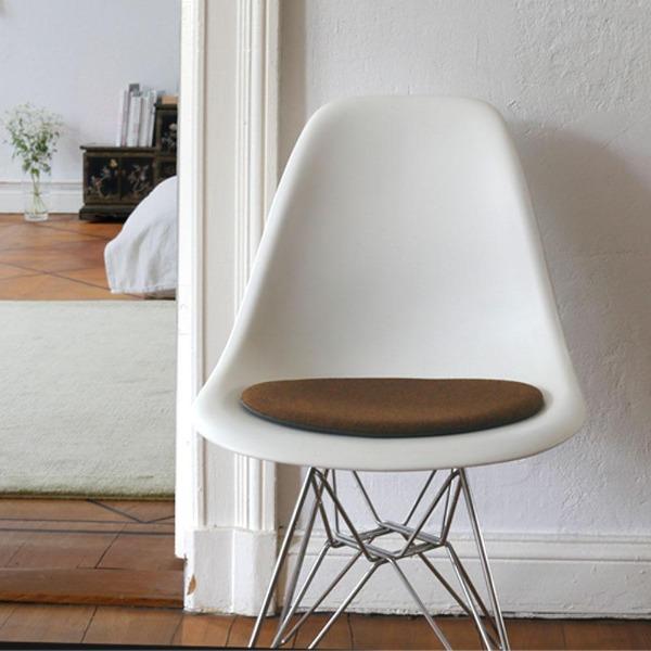 Das foto zeigt einen weissen eames plastic side chair dsr auf dem eine runde sitzauflage der firma discus liegt. Die farbe der sitzauflage ist braun meliert. Der stuhl steht in einem wohnzimmer an der wand.