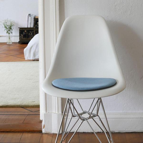 Das foto zeigt einen weissen eames plastic side chair dsr auf dem eine runde sitzauflage der firma discus liegt. Die farbe der sitzauflage ist hellblau meliertt. Der stuhl steht in einem wohnzimmer an der wand.