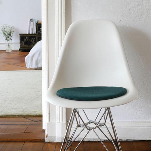 Das foto zeigt einen weissen eames plastic side chair dsr auf dem eine runde sitzauflage der firma discus liegt. Die farbe der sitzauflage ist dunkelgrün meliertt. Der stuhl steht in einem wohnzimmer an der wand.