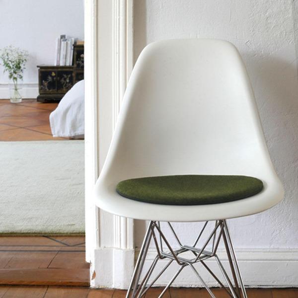 Das foto zeigt einen weissen eames plastic side chair dsr auf dem eine runde sitzauflage der firma discus liegt. Die farbe der sitzauflage ist grün meliert, leichter gelbstich. Der stuhl steht in einem wohnzimmer an der wand.