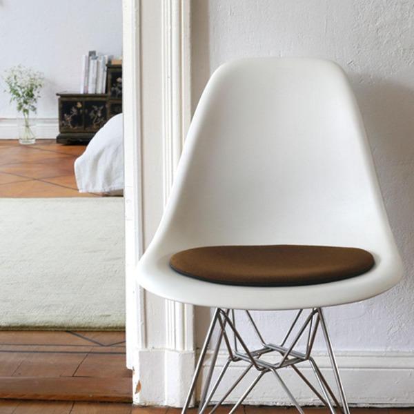 Das foto zeigt einen weissen eames plastic side chair dsr auf dem eine runde sitzauflage der firma discus liegt. Die farbe der sitzauflage ist braun. Der stuhl steht in einem wohnzimmer an der wand.