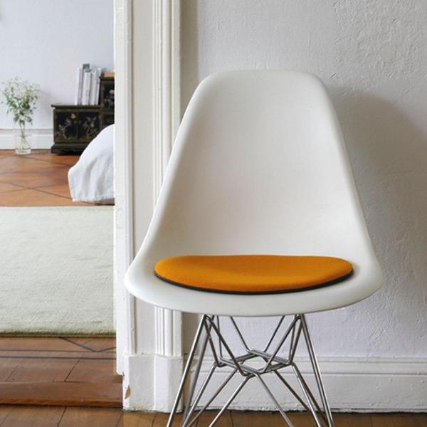 Das foto zeigt einen weissen eames plastic side chair dsr auf dem eine runde sitzauflage der firma discus liegt. Die farbe der sitzauflage ist orange. Der stuhl steht in einem wohnzimmer an der wand.