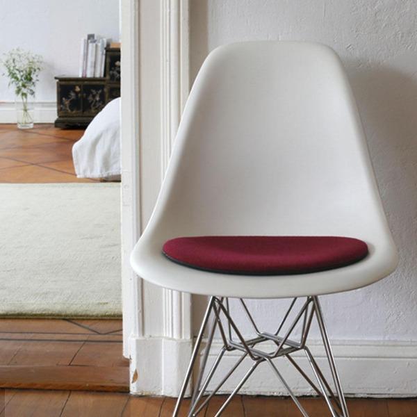 Das foto zeigt einen weissen eames plastic side chair dsr auf dem eine runde sitzauflage der firma discus liegt. Die farbe der sitzauflage ist violett-bordaux meliert. Der stuhl steht in einem wohnzimmer an der wand.