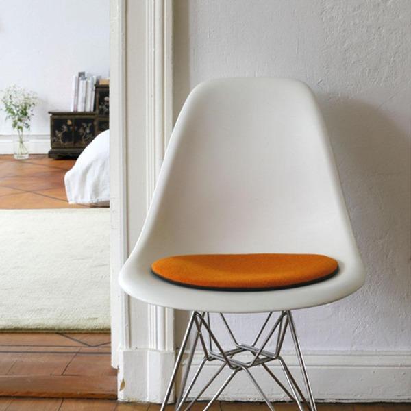 Das foto zeigt einen weissen eames plastic side chair dsr auf dem eine runde sitzauflage der firma discus liegt. Die farbe der sitzauflage ist orange-rot meliert meliert. Der stuhl steht in einem wohnzimmer an der wand.