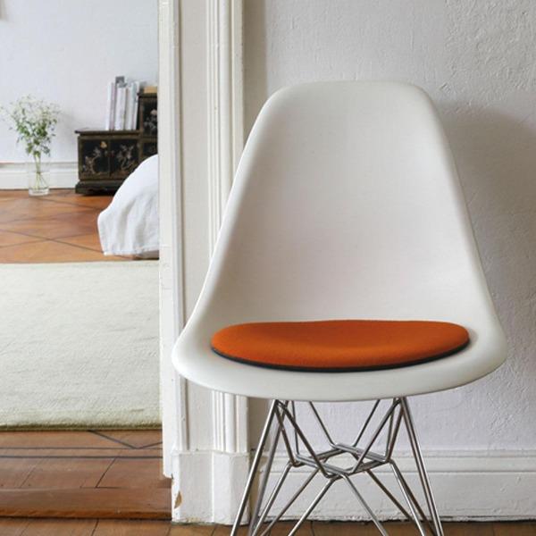 Das foto zeigt einen weissen eames plastic side chair dsr auf dem eine runde sitzauflage der firma discus liegt. Die farbe der sitzauflage ist dunkelorange. Der stuhl steht in einem wohnzimmer an der wand.