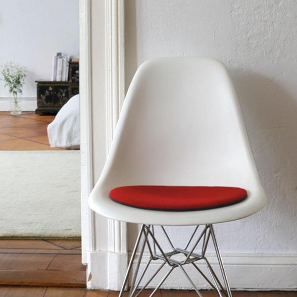  Das foto zeigt einen weissen eames plastic side chair dsr auf dem eine runde sitzauflage der firma discus liegt. Die farbe der sitzauflage ist rot. Der stuhl steht in einem wohnzimmer an der wand.