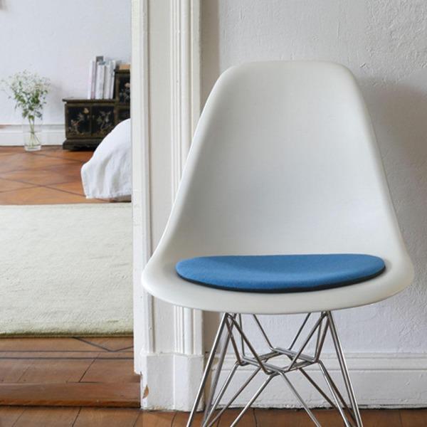 Das foto zeigt einen weissen eames plastic side chair dsr auf dem eine runde sitzauflage der firma discus liegt. Die farbe der sitzauflage ist hellblau. Der stuhl steht in einem wohnzimmer an der wand.