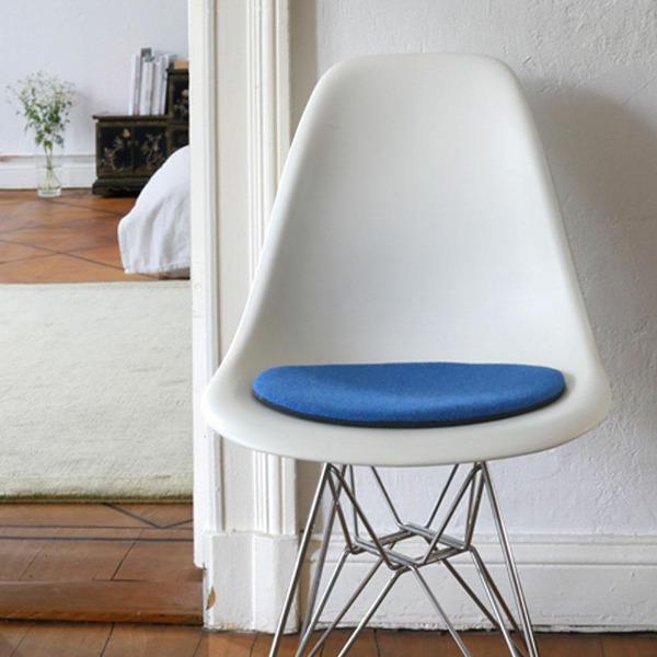 Das foto zeigt einen weissen eames plastic side chair dsr auf dem eine runde sitzauflage der firma discus liegt. Die farbe der sitzauflage ist blau-hellblau meliert. Der stuhl steht in einem wohnzimmer an der wand.