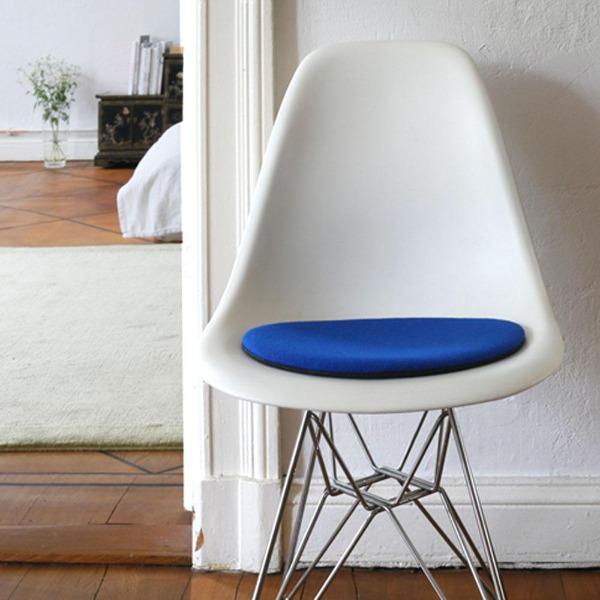 Das foto zeigt einen weissen eames plastic side chair dsr auf dem eine runde sitzauflage der firma discus liegt. Die farbe der sitzauflage ist blau. Der stuhl steht in einem wohnzimmer an der wand.