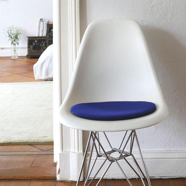 Das foto zeigt einen weissen eames plastic side chair dsr auf dem eine runde sitzauflage der firma discus liegt. Die farbe der sitzauflage ist dunkelblau mit einem leichten rotstich. Der stuhl steht in einem wohnzimmer an der wand.