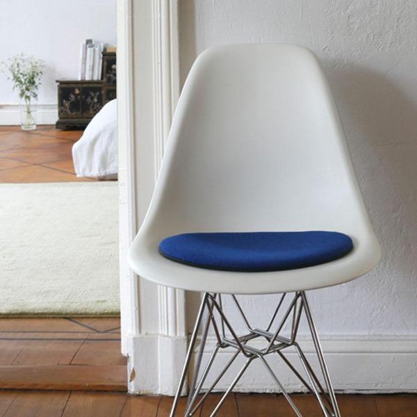 Das foto zeigt einen weissen eames plastic side chair dsr auf dem eine runde sitzauflage der firma discus liegt. Die farbe der sitzauflage ist blau-schwarz meliert. Der stuhl steht in einem wohnzimmer an der wand.