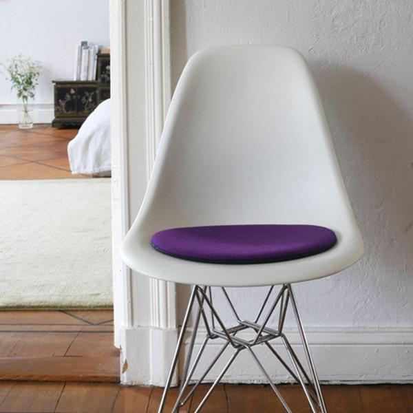 Das foto zeigt einen weissen eames plastic side chair dsr auf dem eine runde sitzauflage der firma discus liegt. Die farbe der sitzauflage ist dunkelblau-violett meliert.. Der stuhl steht in einem wohnzimmer an der wand.