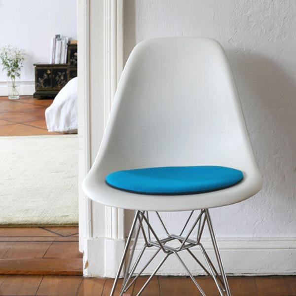 Das foto zeigt einen weissen eames plastic side chair dsr auf dem eine runde sitzauflage der firma discus liegt. Die farbe der sitzauflage ist tuerkis. Der stuhl steht in einem wohnzimmer an der wand.