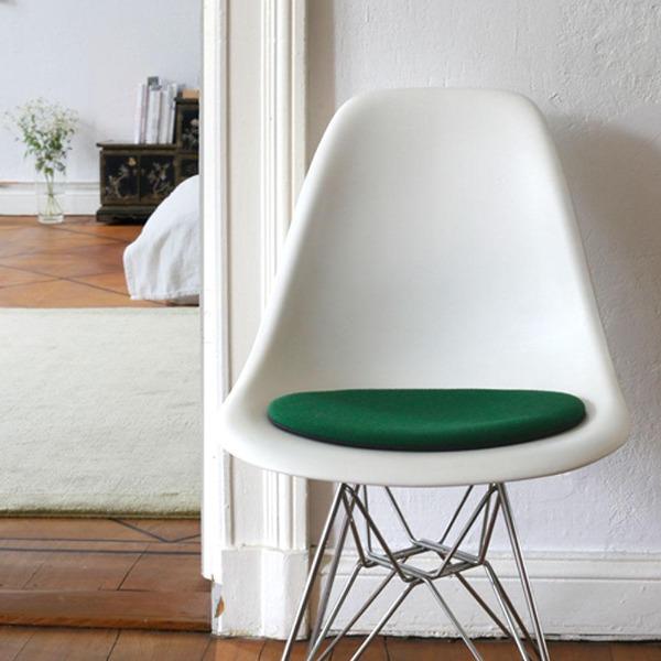 Das foto zeigt einen weissen eames plastic side chair dsr auf dem eine runde sitzauflage der firma discus liegt. Die farbe der sitzauflage ist gruen. Der stuhl steht in einem wohnzimmer an der wand.
