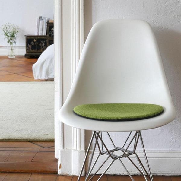 Das foto zeigt einen weissen eames plastic side chair dsr auf dem eine runde sitzauflage der firma discus liegt. Die farbe der sitzauflage ist gelb-gruen meliert. Der stuhl steht in einem wohnzimmer an der wand.