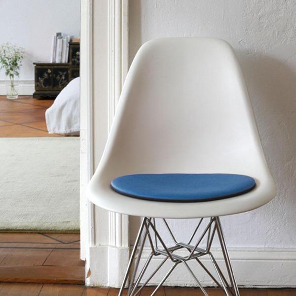 Das foto zeigt einen weissen eames plastic side chair dsr auf dem eine runde sitzauflage der firma discus liegt. Die farbe der sitzauflage ist blau. Der stuhl steht in einem wohnzimmer an der wand.
