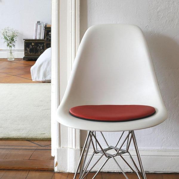Das foto zeigt einen weissen eames plastic side chair dsr auf dem eine runde sitzauflage der firma discus liegt. Die farbe der sitzauflage ist rot. Der stuhl steht in einem wohnzimmer an der wand.