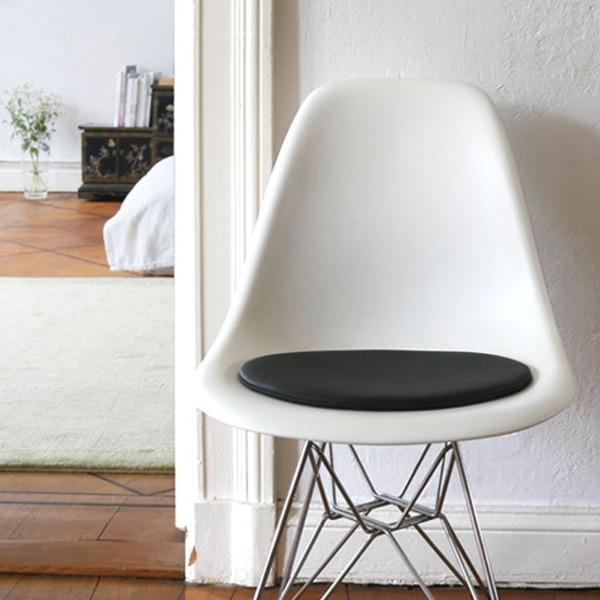 Das foto zeigt einen weissen eames plastic side chair dsr auf dem eine runde sitzauflage der firma discus liegt. Die farbe der sitzauflage ist schwarz. Der stuhl steht in einem wohnzimmer an der wand.