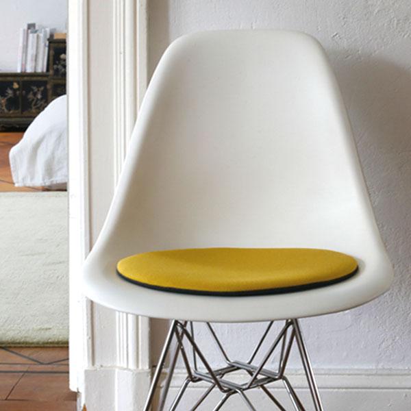 Das foto zeigt einen weissen eames plastic side chair dsr auf dem eine runde sitzauflage der firma discus liegt. Die farbe der sitzauflage ist gelb meliert. Der stuhl steht in einem wohnzimmer an der wand.