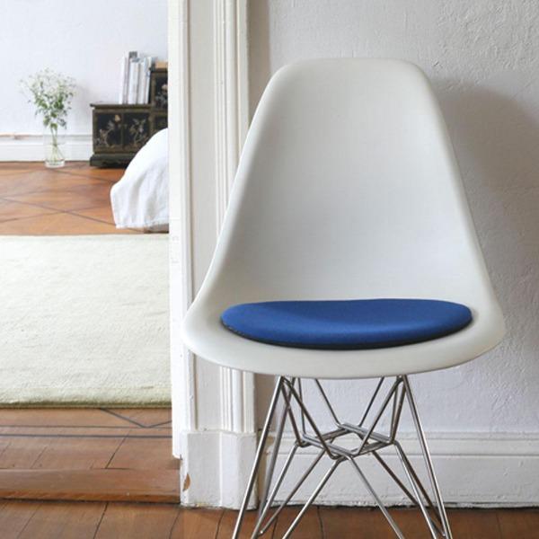 Das foto zeigt einen weissen eames plastic side chair dsr auf dem eine runde sitzauflage der firma discus liegt. Die farbe der sitzauflage ist blau meliert. Der stuhl steht in einem wohnzimmer an der wand.