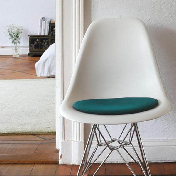 Das foto zeigt einen weissen eames plastic side chair dsr auf dem eine runde sitzauflage der firma discus liegt. Die farbe der sitzauflage ist gruen meliert. Der stuhl steht in einem wohnzimmer an der wand.